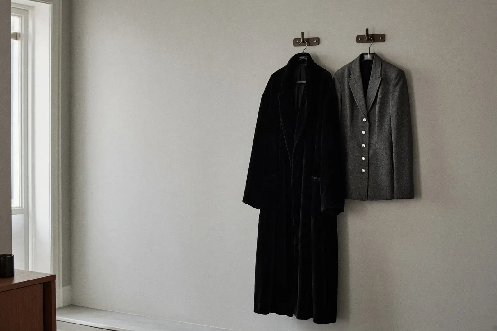 Two jackets on adjacent hooks in a bright modern hallway — one long black velvet, one fitted with narrow lapels and a row of small enamel pins. Bare wall around them.