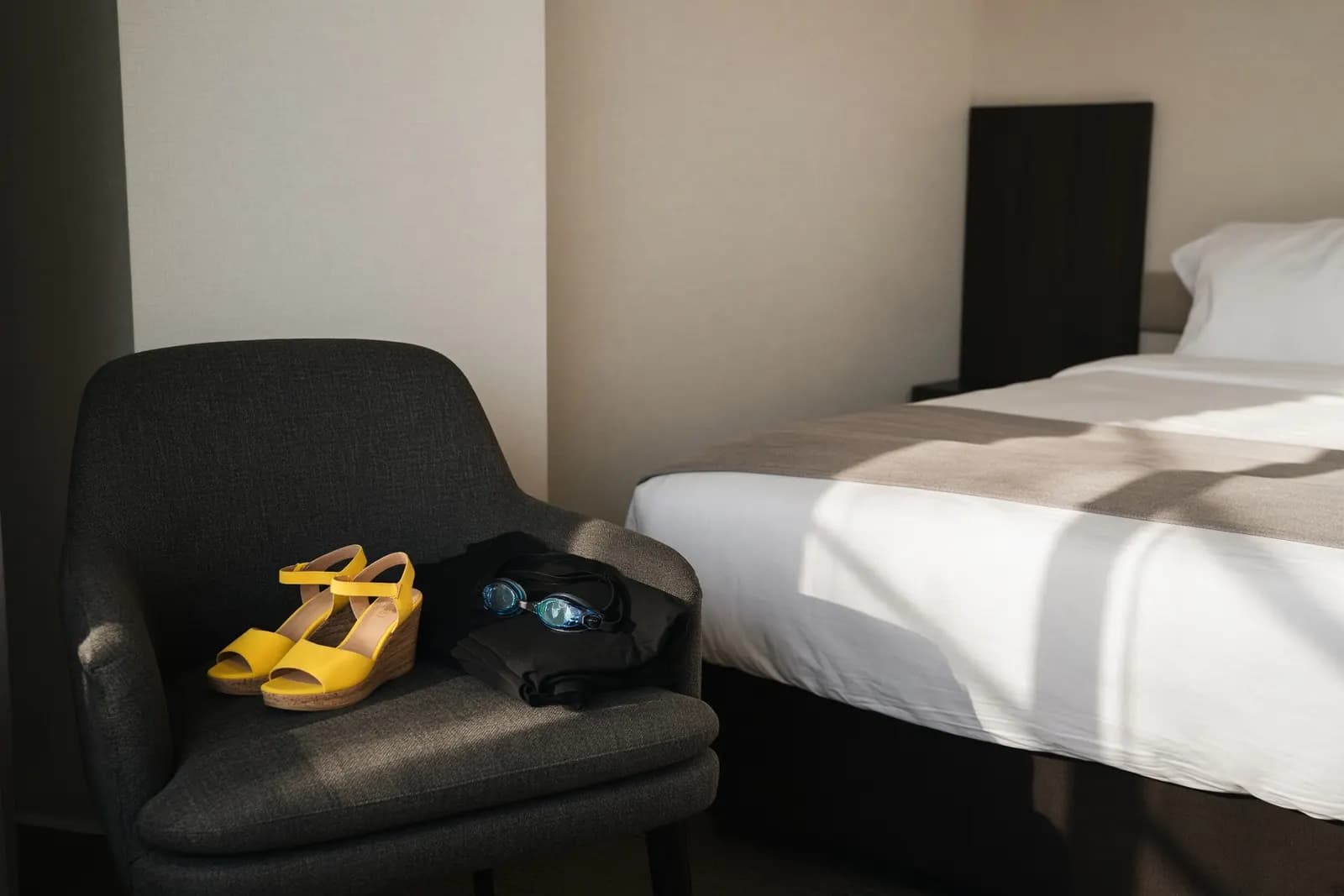 A pair of yellow wedges left on a hotel-room chair beside folded black fabric and swimming goggles, morning light across a made bed.