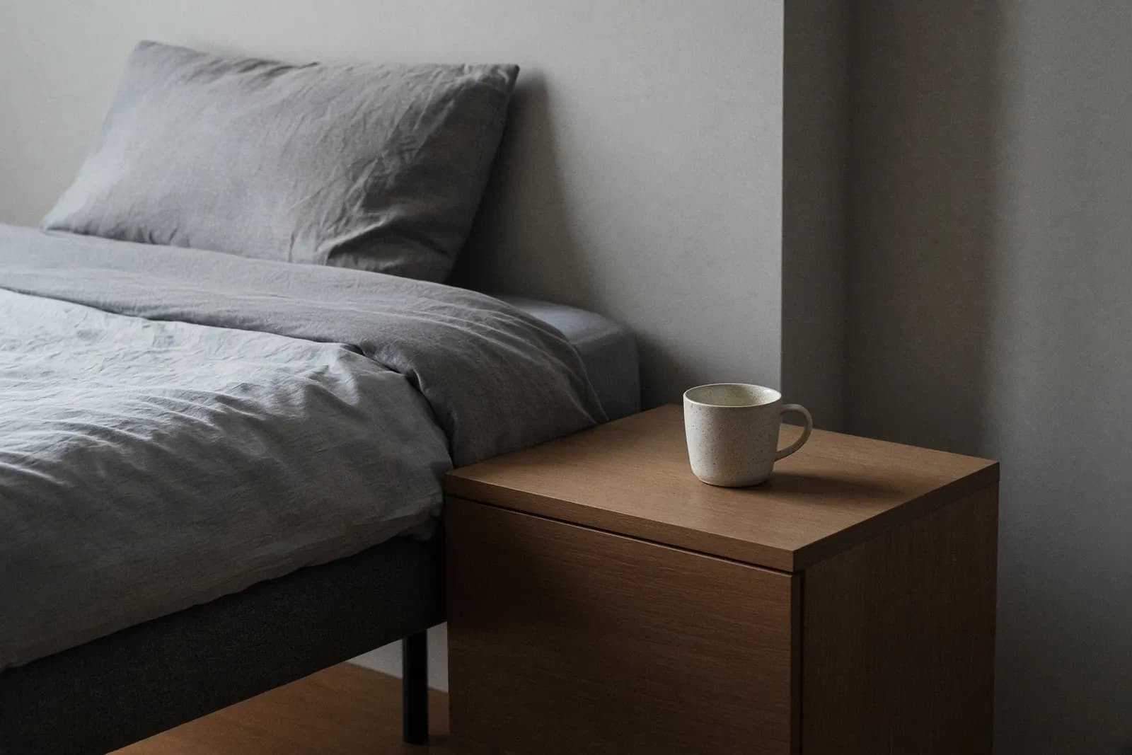 A second mug left on the empty nightstand of a made bed, tea gone cold, in soft morning light.