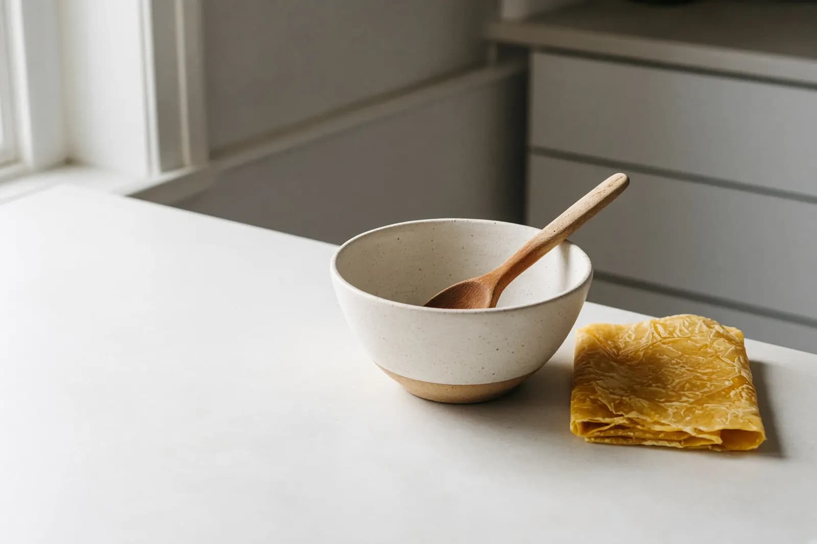 A used wooden spoon left in a mixing bowl beside a folded beeswax wrap on a pale kitchen counter, late-morning light from a side window.